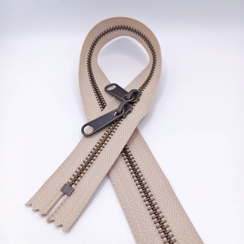 May include: A beige zipper with antique brass sliders. The zipper is open and the sliders are pulled apart.