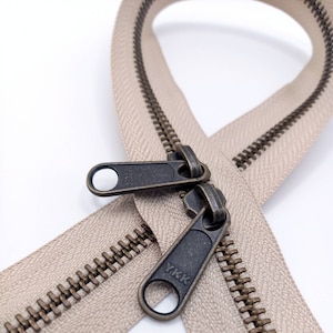 May include: A close-up of a beige zipper with antique brass teeth and a YKK pull tab. The zipper is open and the pull tab is visible.