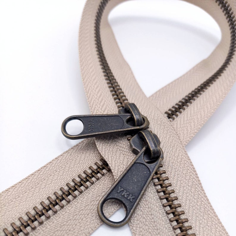 May include: A close-up of a beige zipper with antique brass teeth and a YKK pull tab. The zipper is open and the pull tab is visible.