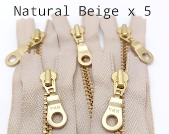 Könnte beinhalten: Fünf beige Reißverschluss-Schieber mit goldenen Zähnen und einem goldenen YKK-Logo. Die Reißverschluss-Schieber sind in einem Stapel angeordnet.