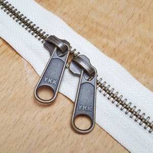 May include: Close-up of a white zipper with two antique brass zipper pulls. The zipper pulls are labelled "YKK".