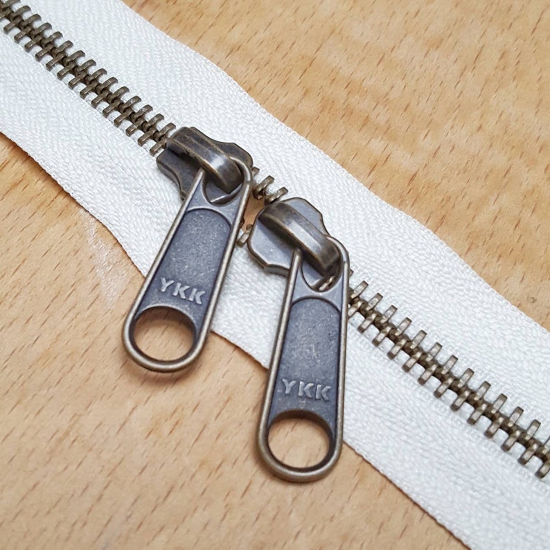 May include: Close-up of a white zipper with two antique brass zipper pulls. The zipper pulls are labelled "YKK".