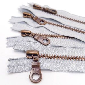 Può includere: Primo piano di cinque cerniere argentate con tiretti in ottone. Le cerniere sono tutte del marchio YKK e hanno una finitura argentata. I tiretti sono tutti in ottone e hanno un anello nella parte superiore.