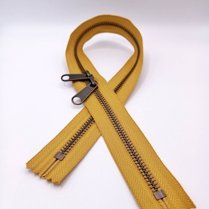 May include: A yellow zipper with antique brass sliders. The zipper is laid out in a figure of eight shape.