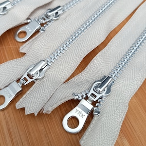 Può includere: Primo piano di quattro nastri di cerniera beige con denti e cursori in metallo argentato. I cursori hanno un anello in metallo argentato e il testo "YKK" è stampato sul cursore.