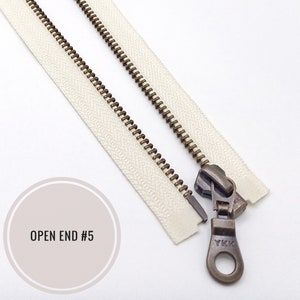 Pode incluir: Um zíper branco com um cursor e um puxador de latão. O zíper está aberto e tem a etiqueta "OPEN END #5".