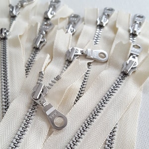 Könnte beinhalten: Nahaufnahme mehrerer cremefarbener Reißverschlüsse mit silbernen Metallschiebern. Die Reißverschlüsse sind in verschiedenen Winkeln angeordnet und zeigen die Metallzähne und die Marke YKK auf den Schiebern. Der Stoff ist in einem weichen Off-White-Ton gehalten.