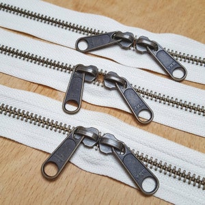 May include: Three white zipper tapes with antique brass metal zipper pulls. The zipper pulls are double ended and have the text "YKK" on them.