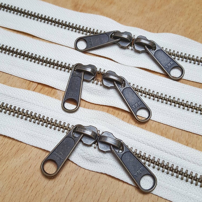 May include: Three white zipper tapes with antique brass metal zipper pulls. The zipper pulls are double ended and have the text "YKK" on them.