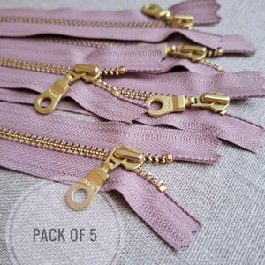 May include: Close-up of five mauve-colored zippers with gold-toned metal teeth and pulls. The zippers are arranged in a slightly overlapping pattern. The text "PACK OF 5" is visible in a circle.