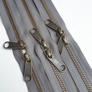 May include: Three grey zipper tapes with antique brass zipper pulls. The zipper pulls are labelled "YKK".