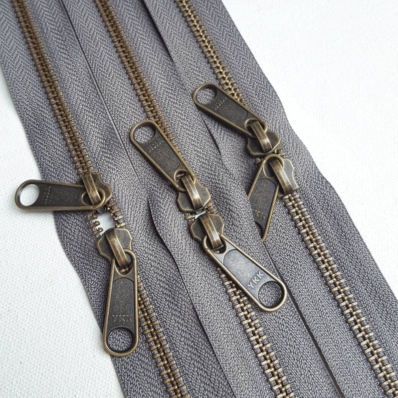 May include: Three grey zipper tapes with antique brass zipper pulls. The zipper pulls are labelled "YKK".