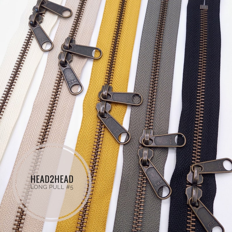 May include: Five zipper tapes with antique brass sliders and pulls. The tapes are in white, beige, yellow, grey, and black. The zipper pulls are labelled "YKK" and say "HEAD2HEAD LONG PULL #5".