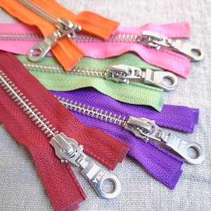 Può includere: Assortimento di nastri per cerniere colorati con cursori in metallo argentato. Le cerniere sono disposte a forma di arcobaleno, con nastri rossi, viola, verdi, rosa e arancioni. Tutte le cerniere sono chiuse e hanno il nome del marchio YKK sul cursore.