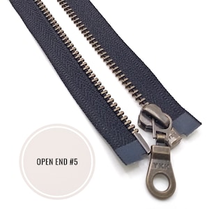 Pode incluir: Um zíper preto com um cursor e um puxador de latão. O zíper está aberto e o puxador é etiquetado com "YKK". O zíper é etiquetado com "OPEN END #5".