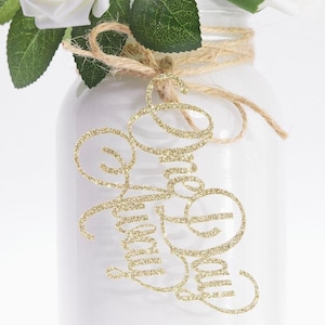 May include: A white painted mason jar with a twine bow and gold glitter script that reads "Love You". The jar holds white roses with green leaves. A decorative floral arrangement.