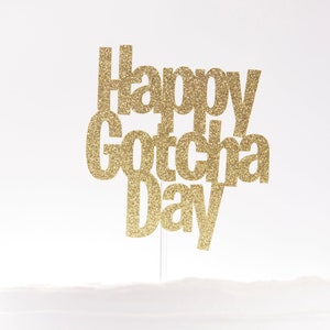 May include: A gold glitter cake topper featuring the text Happy Gotcha Day in a stacked sans-serif font. The topper is attached to a thin, clear vertical stick, designed to be inserted into a cake for celebratory display and decoration.