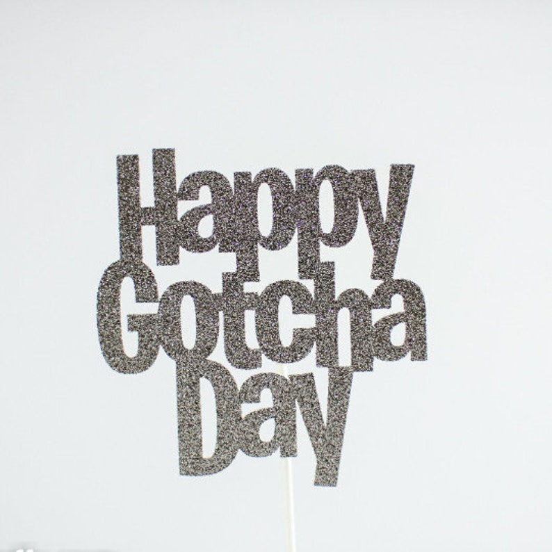 May include: A cake topper featuring the text Happy Gotcha Day in a bold, sans-serif font. The letters are covered in a dark silver glitter finish, creating a textured, sparkling appearance. A thin, white support stick is attached to the bottom.