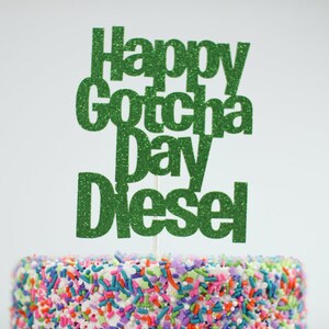 May include: A green glitter cake topper featuring the text Happy Gotcha Day Diesel in a bold sans-serif font. The topper is attached to a thin white stick and is positioned above a cake covered in colorful, multi-shaped sprinkles.