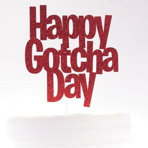 May include: A red glitter cake topper featuring the text Happy Gotcha Day in a bold, sans-serif font. The lettering is attached to a thin, clear vertical stick, designed to be inserted into a cake for celebratory display purposes.