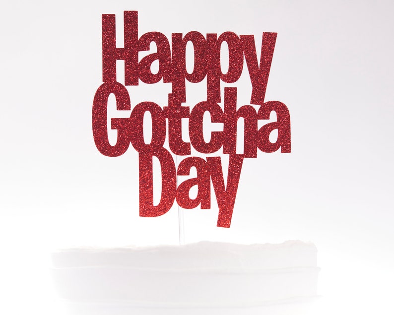 May include: A red glitter cake topper featuring the text Happy Gotcha Day in a bold, sans-serif font. The lettering is attached to a thin, clear vertical stick, designed to be inserted into a cake for celebratory display purposes.