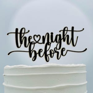 May include: A black cake topper with the words "the night before" in a cursive font. The topper is placed on top of a white frosted cake. The heart symbol replaces the letter "o" in the word "the".