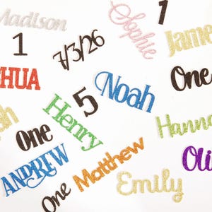 May include: A collection of glittery cardstock cake toppers featuring various names including Madison, Sophie, James, Joshua, Noah, Elijah, Henry, Hannah, Andrew, Matthew, Olivia, and Emily, along with the numbers 1, 5, and the date 7/3/26.