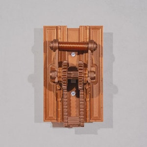 May include: A bronze-colored, rectangular wall-mounted decorative piece with a mechanical gear design. The piece features a central handle, two gears, and a series of vertical and horizontal lines, all in a metallic finish. The design is symmetrical and detailed.