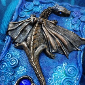 Puede incluir: Una escultura de drag&oacute;n de bronce con alas azules y una gema azul en la cola, sobre un fondo azul con remolinos blancos.