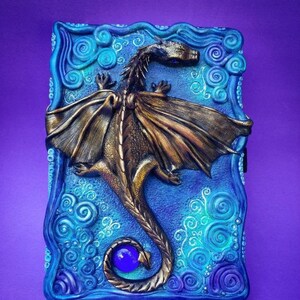 Puede incluir: Un drag&oacute;n azul y dorado con las alas extendidas est&aacute; esculpido en la portada de un diario. La cola del drag&oacute;n se enrosca alrededor de una gema azul. El fondo es un patr&oacute;n azul y verde en espiral.