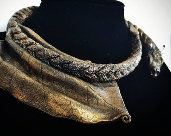 May include: A gold-toned necklace with a leaf-shaped pendant. The necklace is made of braided metal and has a textured surface.