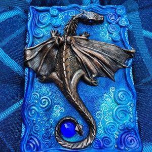 Puede incluir: Una caja de cer&aacute;mica azul y blanca con un dise&ntilde;o de drag&oacute;n esculpido. El drag&oacute;n tiene un ojo azul y una gema azul en su cola. La caja est&aacute; decorada con patrones en espiral.