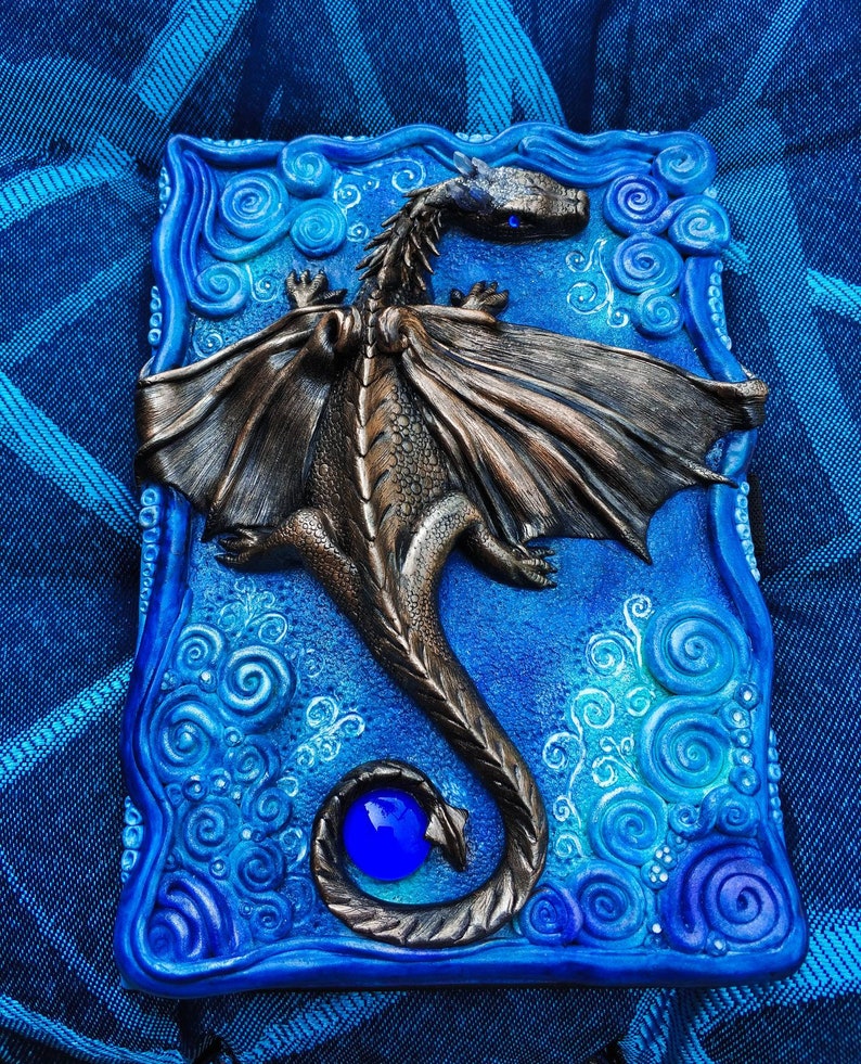 Puede incluir: Una caja de cer&aacute;mica azul y blanca con un dise&ntilde;o de drag&oacute;n esculpido. El drag&oacute;n tiene un ojo azul y una gema azul en su cola. La caja est&aacute; decorada con patrones en espiral.