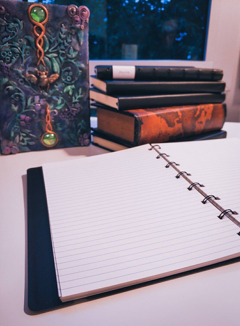 Puede incluir: Un cuaderno abierto de espiral con p&aacute;ginas rayadas y una cubierta negra. El cuaderno est&aacute; sobre una superficie blanca con una pila de libros y una caja decorativa con gemas verdes y dise&ntilde;os florales.