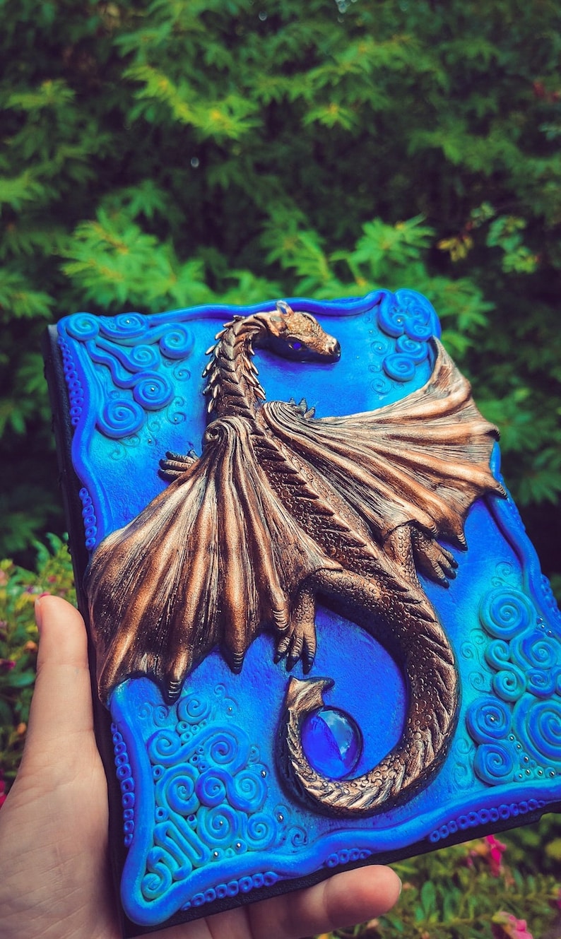 Puede incluir: Un diario azul y verde con un dise&ntilde;o de drag&oacute;n esculpido. El drag&oacute;n est&aacute; hecho de arcilla marr&oacute;n y tiene una gema azul en su cola.