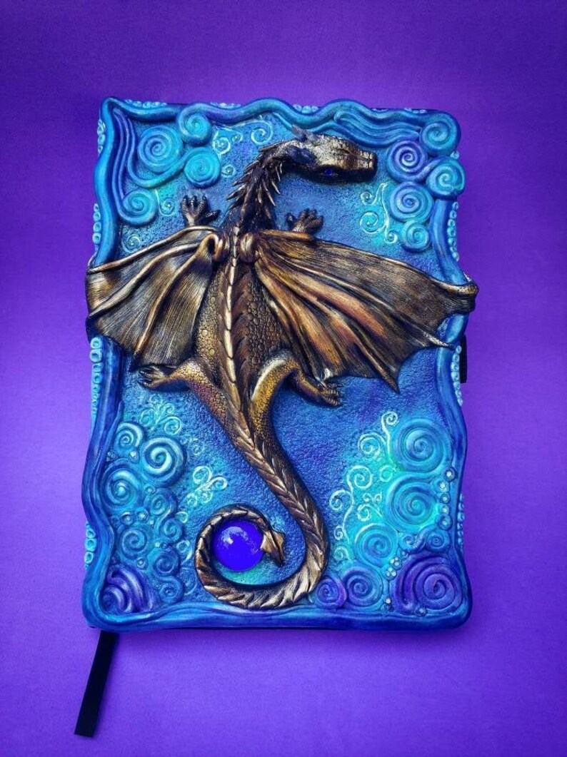 Puede incluir: Un drag&oacute;n azul y dorado con las alas extendidas est&aacute; esculpido en la portada de un diario. La cola del drag&oacute;n se enrosca alrededor de una gema azul. El fondo es un patr&oacute;n azul y verde en espiral.