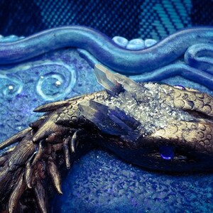 Puede incluir: Primer plano de una escultura de drag&oacute;n dorada con una corona de cristal, sobre un fondo azul y blanco. El ojo del drag&oacute;n es una piedra preciosa azul.