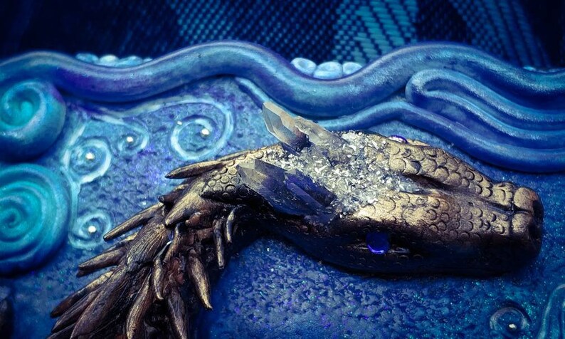 Puede incluir: Primer plano de una escultura de drag&oacute;n dorada con una corona de cristal, sobre un fondo azul y blanco. El ojo del drag&oacute;n es una piedra preciosa azul.
