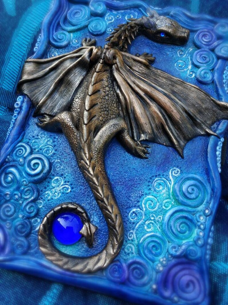 Puede incluir: Una escultura de drag&oacute;n de bronce con alas azules y una gema azul en la cola, sobre un fondo azul con remolinos blancos.