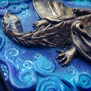 Puede incluir: Primer plano de una escultura de drag&oacute;n de bronce con escamas y garras detalladas, descansando sobre un fondo azul y blanco con patrones en espiral.