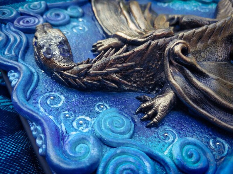 Puede incluir: Primer plano de una escultura de drag&oacute;n de bronce con escamas y garras detalladas, descansando sobre un fondo azul y blanco con patrones en espiral.