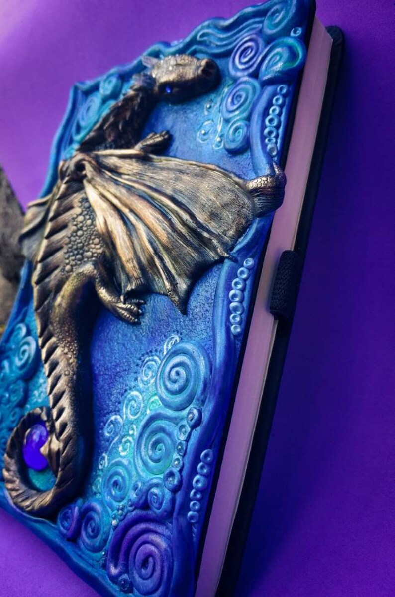 Puede incluir: Un diario con una portada esculpida que representa un drag&oacute;n azul y dorado. El drag&oacute;n tiene un cuerpo azul con escamas y alas dor&eacute;es. La portada del diario est&aacute; decorada con espirales y puntos en tonos azules y morados.