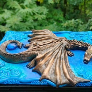 Puede incluir: Una caja decorativa azul y verde con un drag&oacute;n de bronce esculpido en la tapa. El drag&oacute;n tiene una gema azul en su cola y sus alas est&aacute;n extendidas.