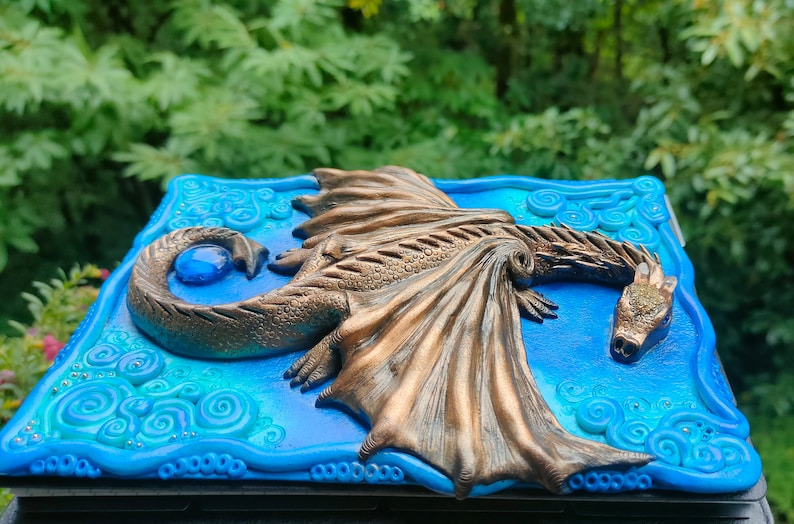 Puede incluir: Una caja decorativa azul y verde con un drag&oacute;n de bronce esculpido en la tapa. El drag&oacute;n tiene una gema azul en su cola y sus alas est&aacute;n extendidas.