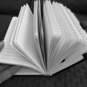 Puede incluir: Una imagen en blanco y negro de un cuaderno con p&aacute;ginas rayadas abiertas en una p&aacute;gina en blanco. El cuaderno est&aacute; acostado sobre una superficie texturizada.