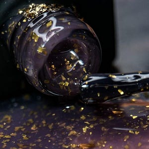 May include: Close-up of a nail polish bottle with a dark purple hue and gold flakes. The applicator brush is extended, and the polish is partially applied. The image highlights the texture and colour of the nail polish.