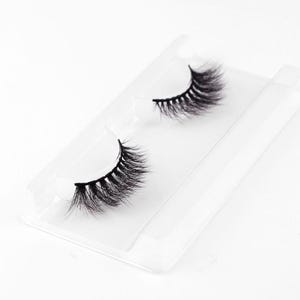 Könnte beinhalten: Ein Paar schwarze falsche Wimpern, präsentiert auf einer transparenten Kunststoffschale. Die Wimpern sind dicht und voll, mit einem natürlichen Schwung. Die Schale ist rechteckig mit einer gebogenen Oberseite, um die Wimpern sicher zu halten.
