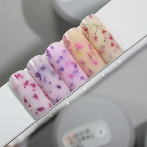 May include: Five rectangular nail tips arranged in a row on a white display stand, numbered one through five. Each tip features a translucent base color ranging from light pink to cream, embedded with small, colorful dried flower petals throughout.