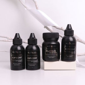 Peut inclure: Quatre flacons noirs de vernis à ongles avec des lettres dorées. Les flacons sont étiquetés "KOMILFO" et contiennent "TOP COAT" et "RUBBER BASE COAT".