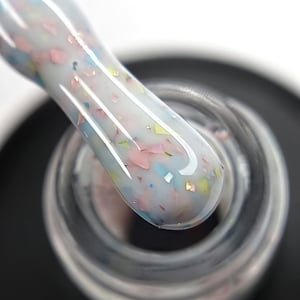 May include: Close-up of a nail polish brush with a white base and colourful confetti-like pieces. The brush is dipped into a clear bottle of nail polish. The confetti pieces are pink, blue, yellow, and iridescent.